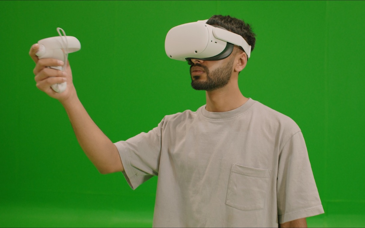 young man in grey t-shirt wearing a meta quest vr headset and holding motion tracking hand device in front of green screen